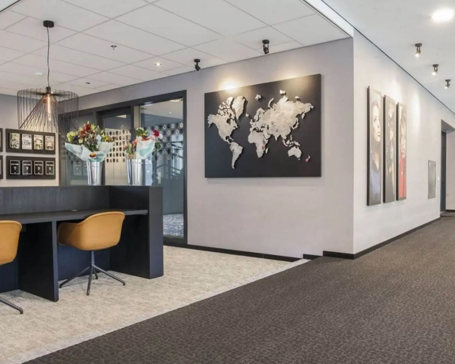 Moderne kantoorruimte met wanddecoratie waaronder een wereldkaart en portretfoto's, en een werkplek met bloemen op het bureau.