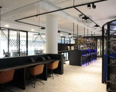 Moderne kantoorlounge met werkplekken, een bar met blauwe verlichting en glazen vergaderruimtes op de achtergrond.