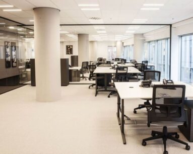 Moderne, open kantoorruimte met meerdere bureaus, bureaustoelen en grote ramen voor natuurlijk licht.
