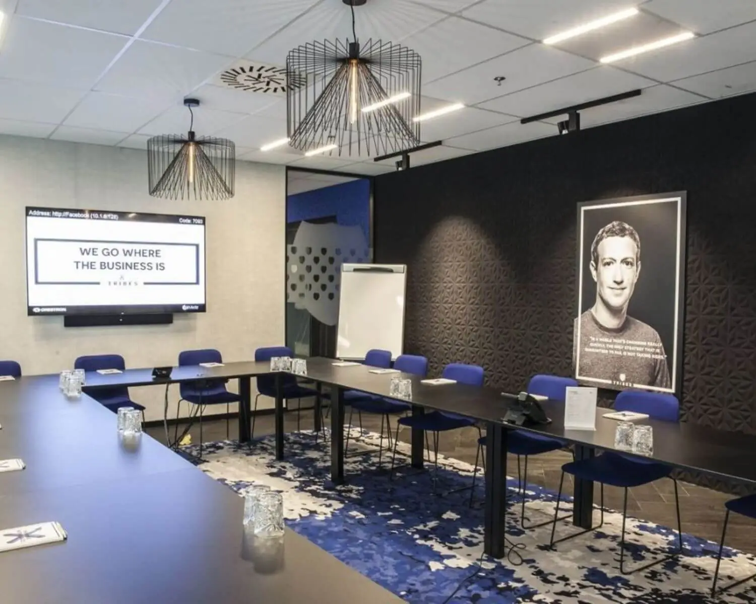 Moderne vergaderruimte met een U-vormige tafelopstelling, blauwe stoelen, een scherm met de tekst "We go where the business is" en een grote zwart-wit portretfoto aan de muur.