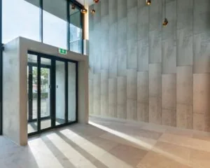 Moderne entreehal met glazen schuifdeuren, hoge plafonds en een wand van betonpanelen.