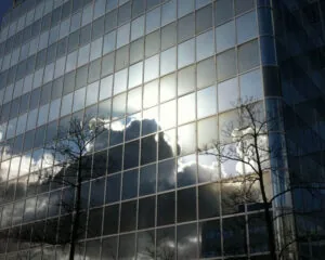 Reflecterende glazen gevel van een kantoorgebouw met weerkaatsing van wolken, zonlicht en silhouetten van bomen.