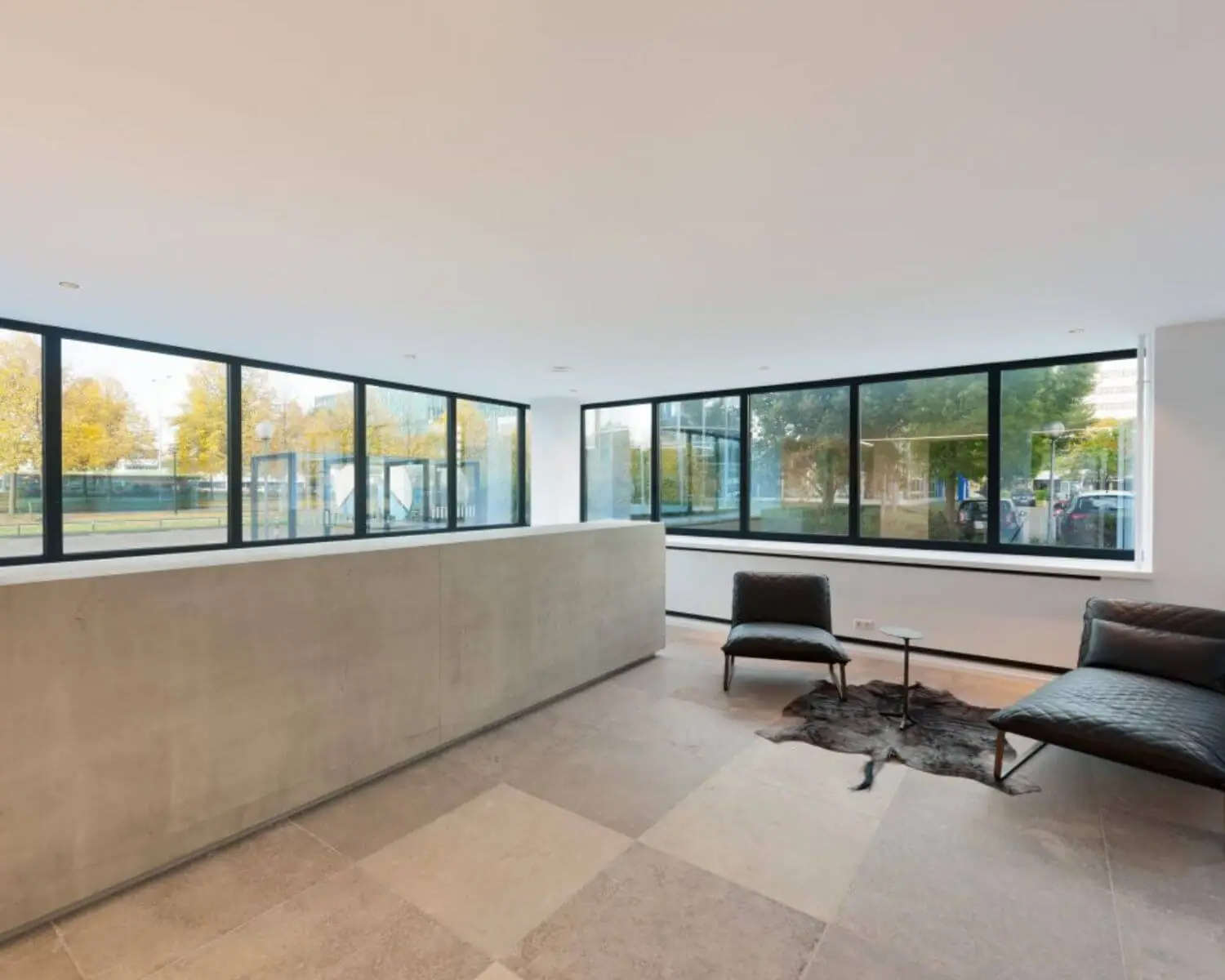 Moderne wachtruimte met grote ramen, betonnen balie, leren meubels en uitzicht op een stedelijke omgeving.