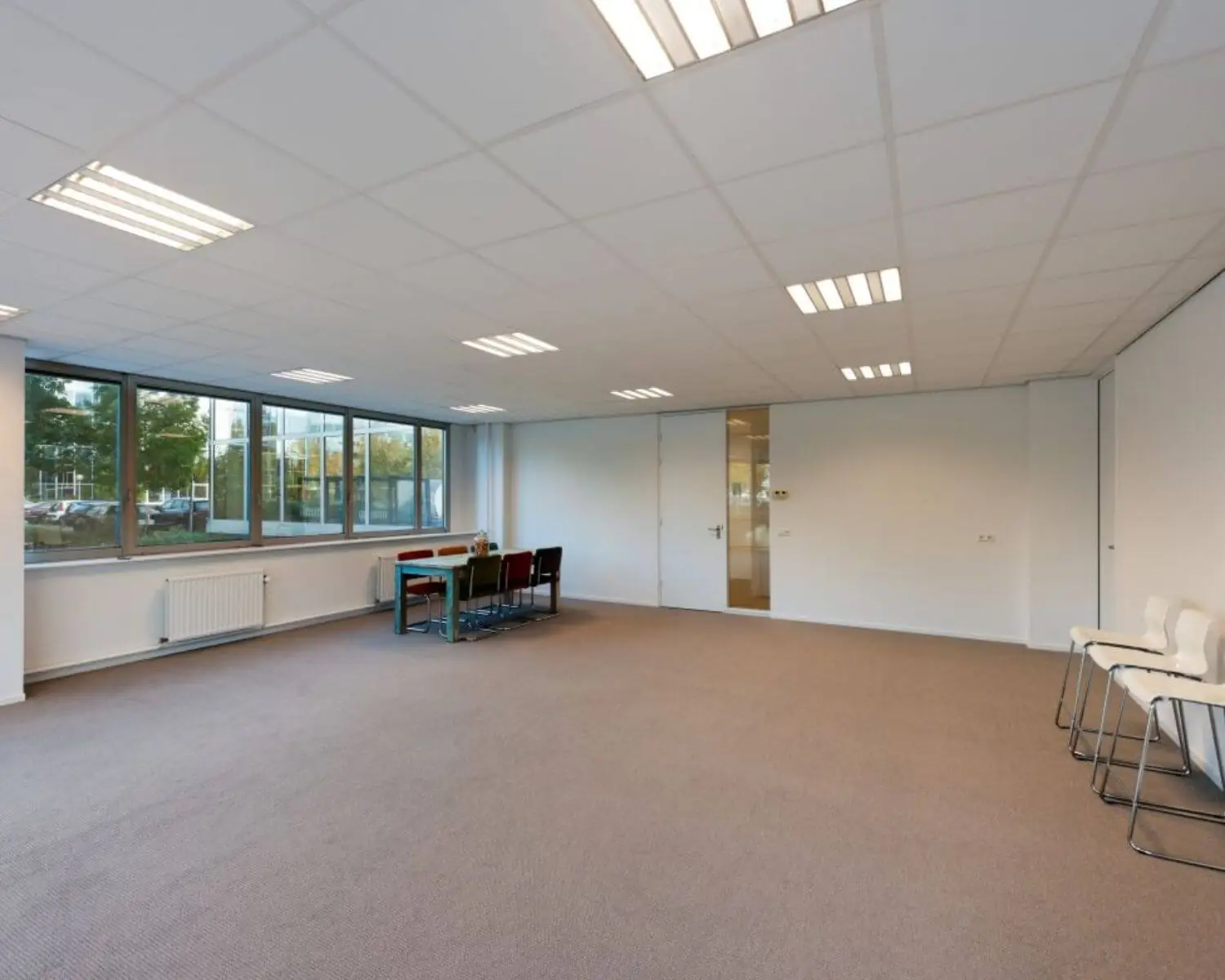 Ruime, lichte kantoorkamer met grote ramen, systeemplafond en een vergadertafel met stoelen.