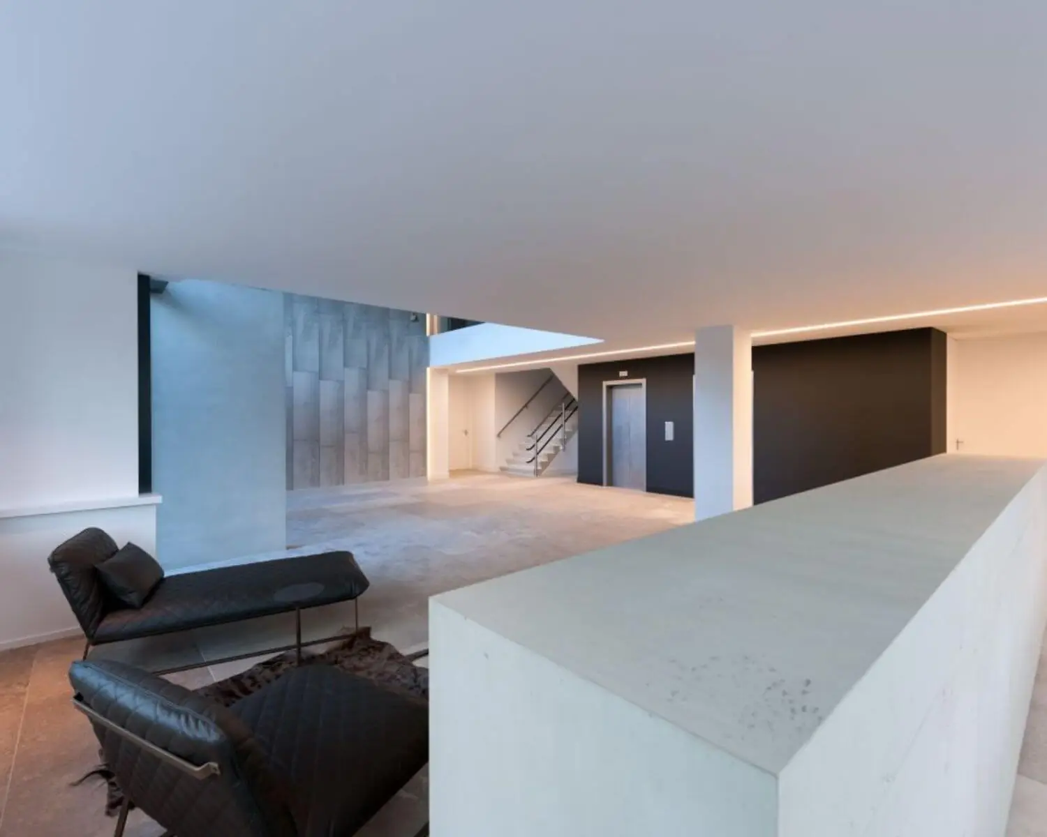 Minimalistisch en modern interieur met loungestoelen, een betonnen trap en een lift in een open ruimte met strakke lijnen.