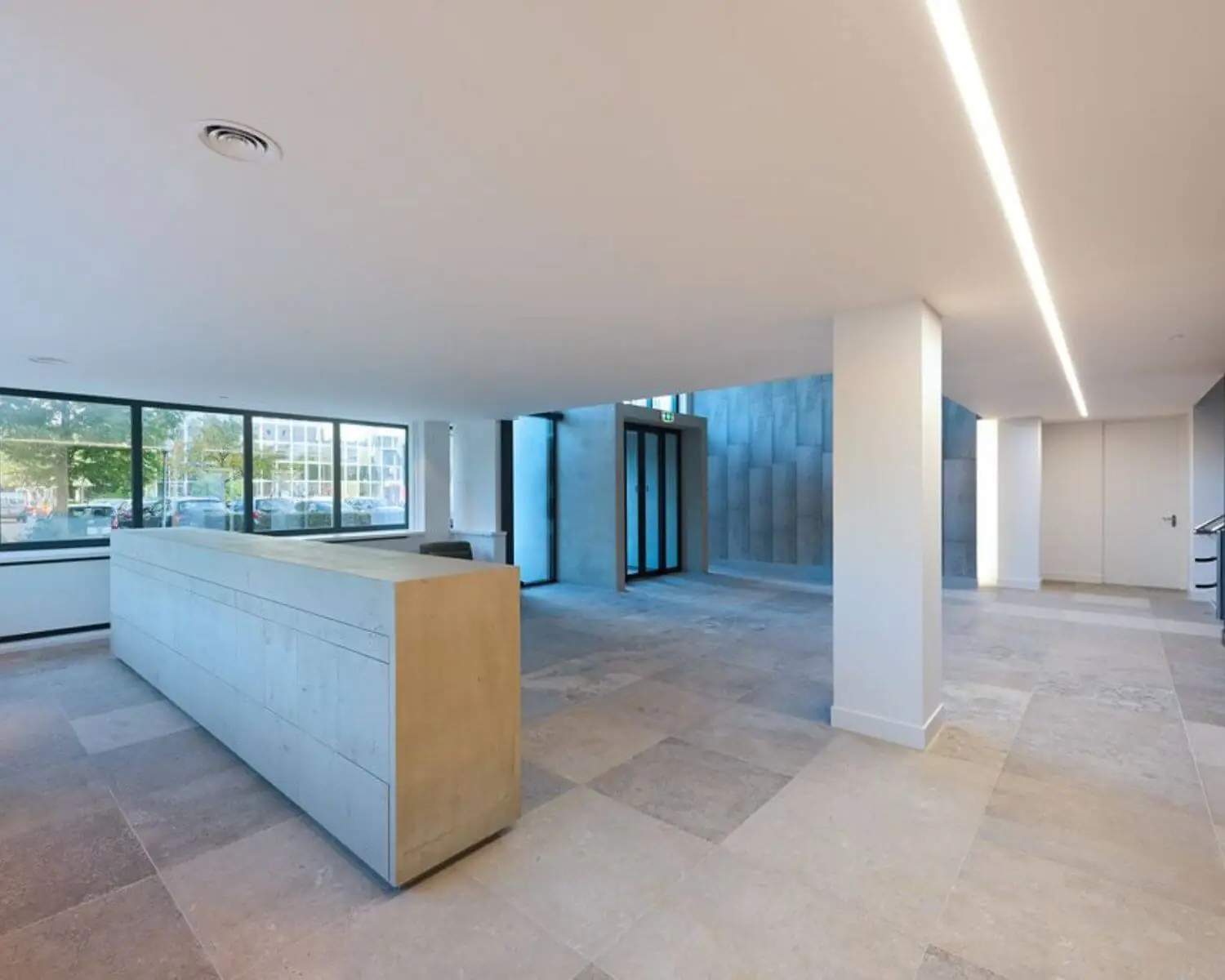 Moderne, lichte entreehal met een betonnen receptiebalie, grote raampartijen en een strakke, minimalistische afwerking.
