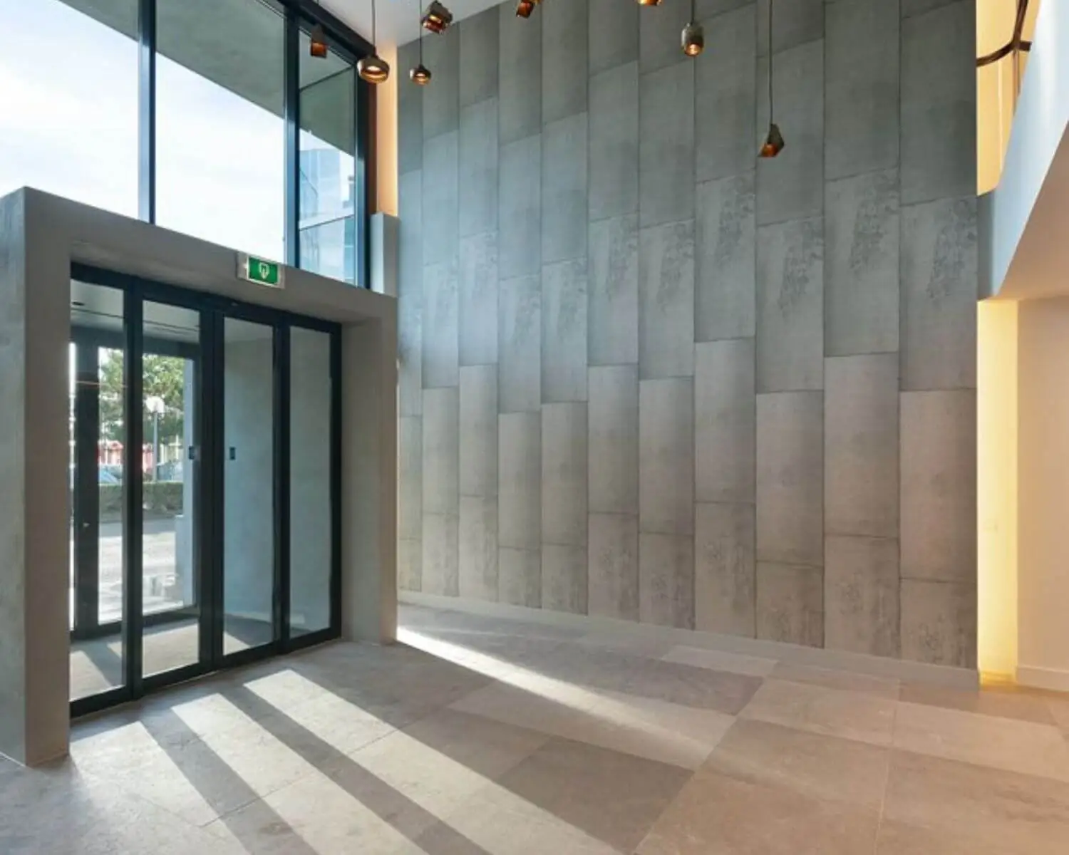 Moderne entreehal met hoge plafonds, betonnen wandpanelen en een glazen schuifdeur.
