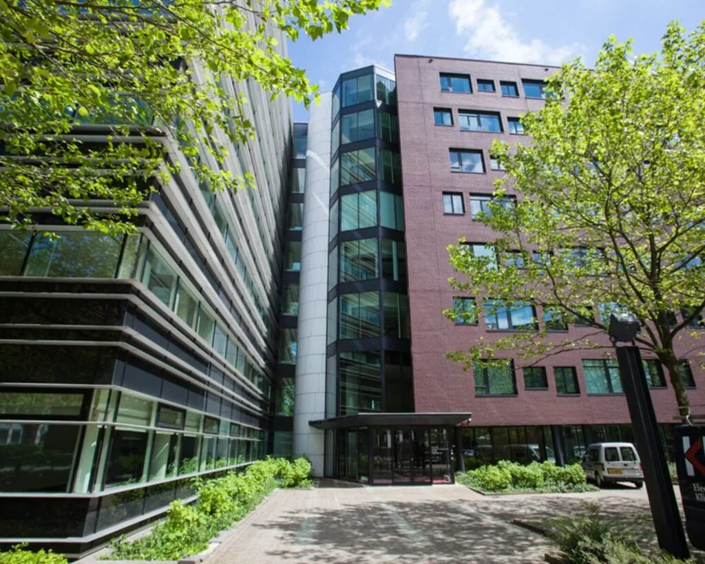 Vooraanzicht van een modern kantoorgebouw met glas- en baksteenfacades, omringd door groen.
