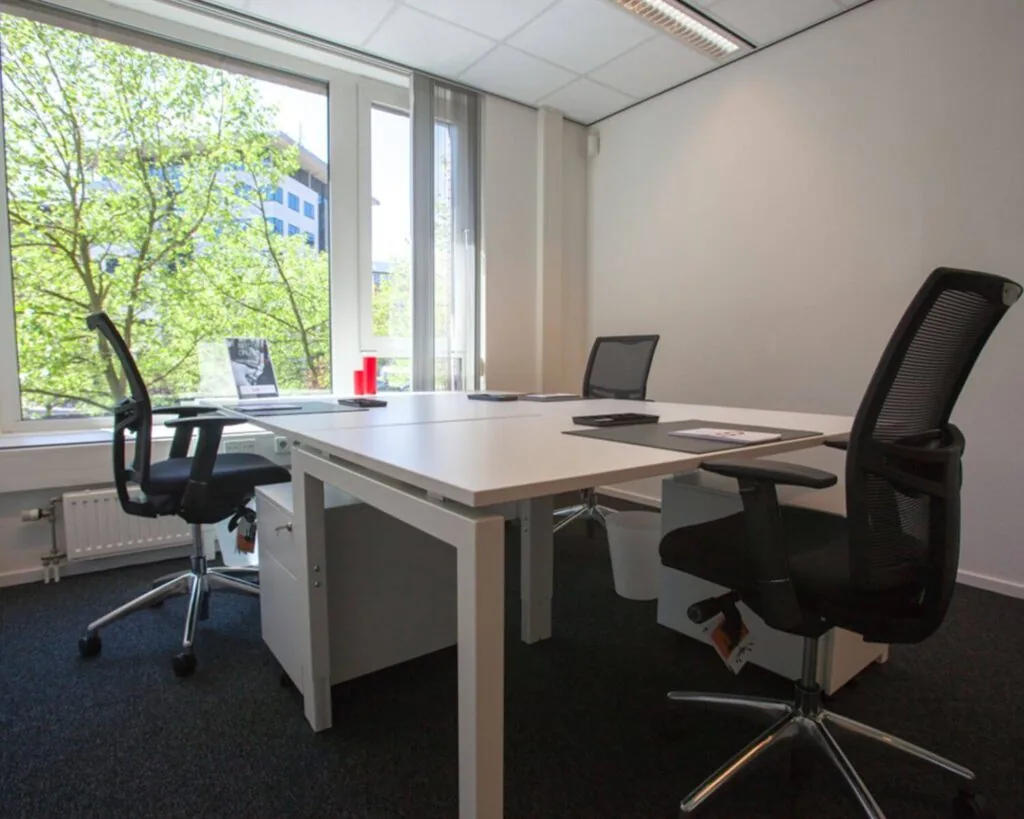 Modern kantoor met vier bureaustoelen en een groot raam met uitzicht op groene bomen.