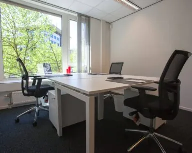 Modern kantoor met vier bureaustoelen en een groot raam met uitzicht op groene bomen.