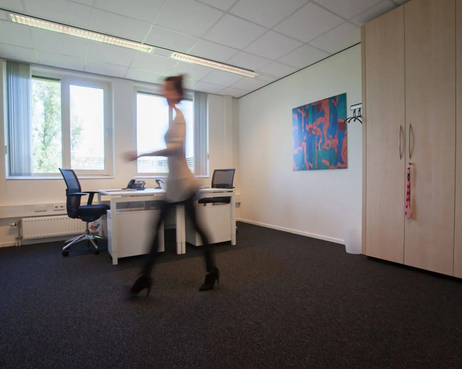 Een vrouw loopt door een modern kantoor met twee bureaus, bureaustoelen en kleurrijke schilderkunst aan de muur.