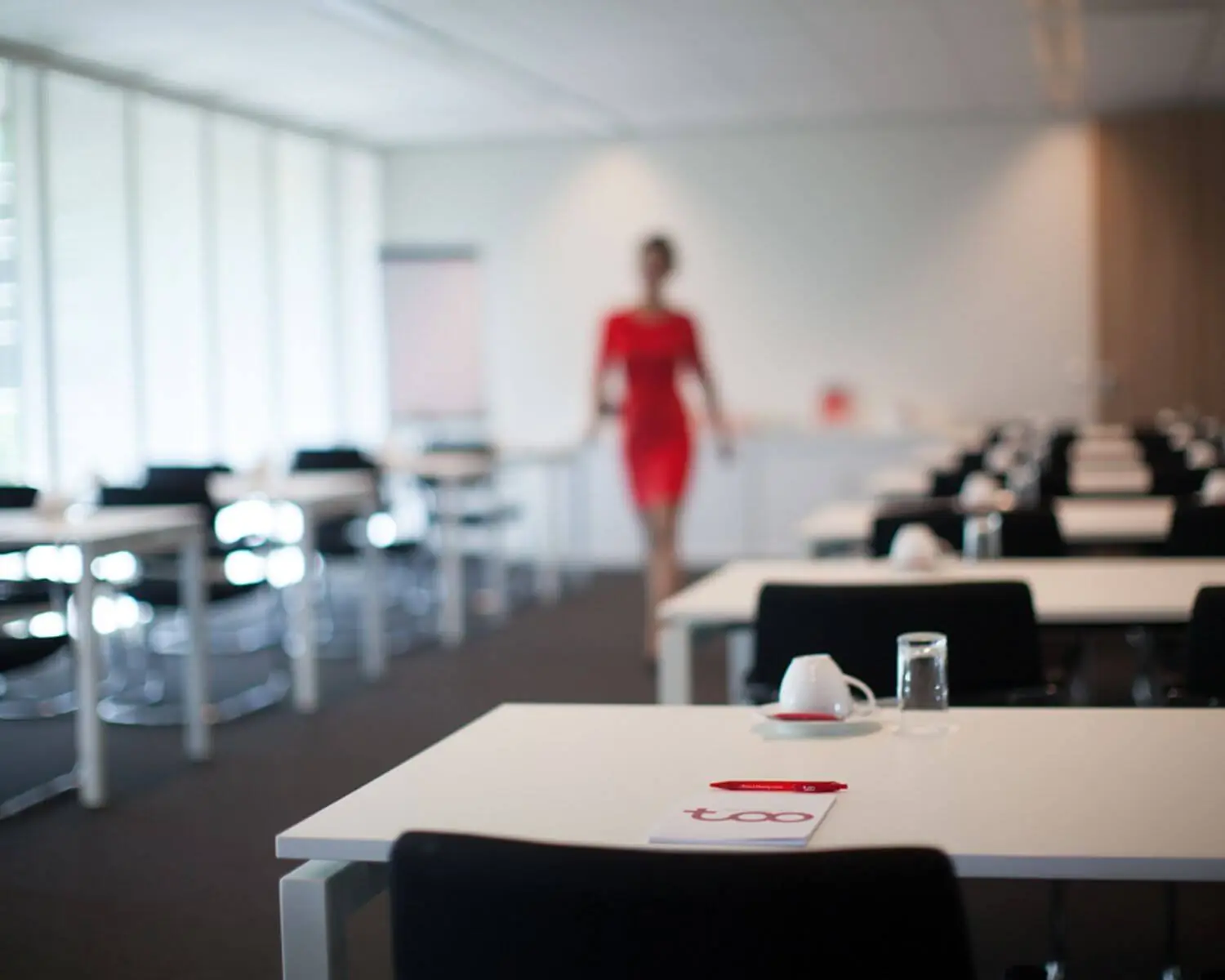 Een moderne vergaderruimte met opgestelde tafels, een notitieboekje en een vrouw in een rode jurk op de achtergrond.