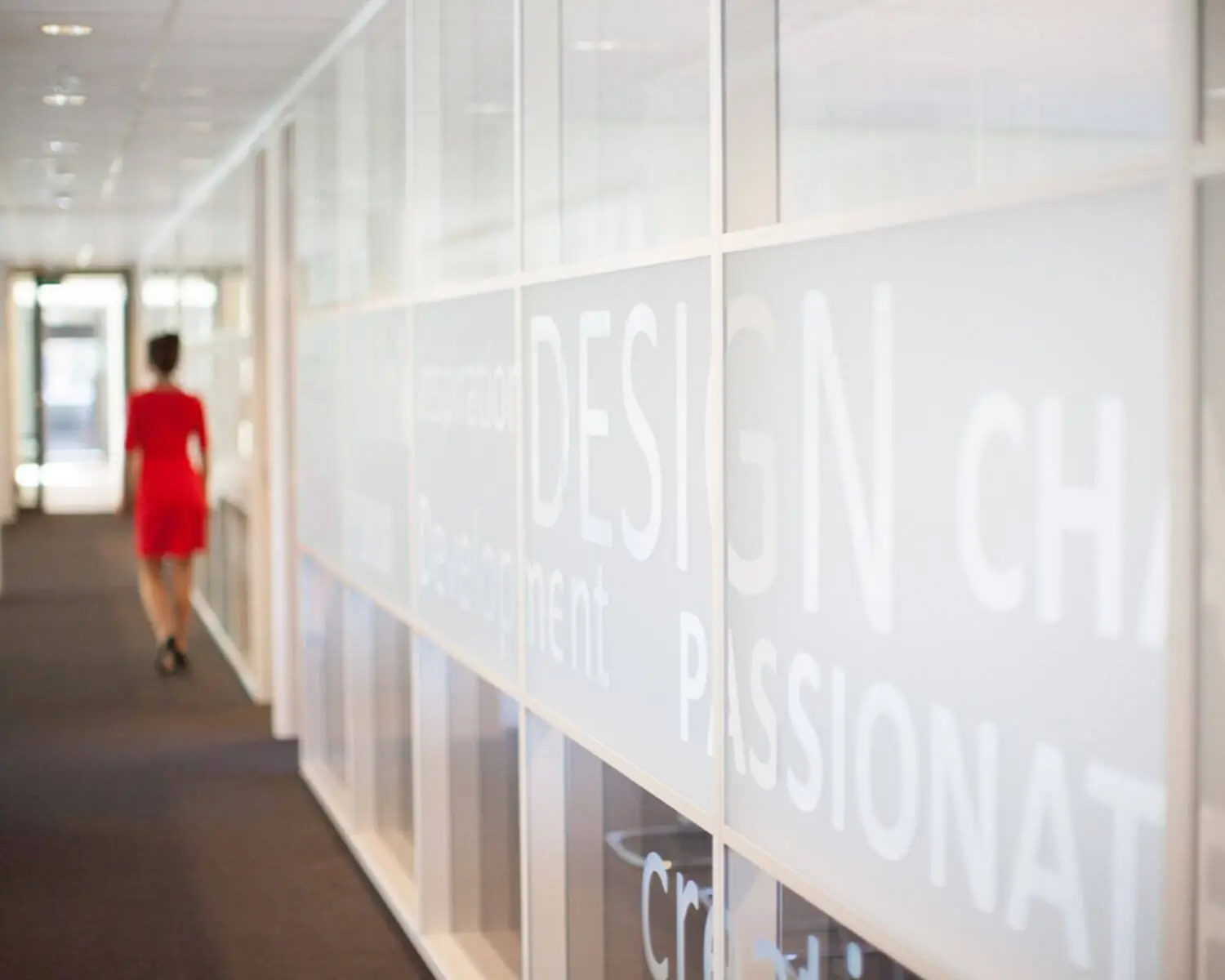 Vrouw in rode jurk loopt door een modern kantoor met glazen wand vol inspirerende woorden zoals "DESIGN" en "PASSIONATE".