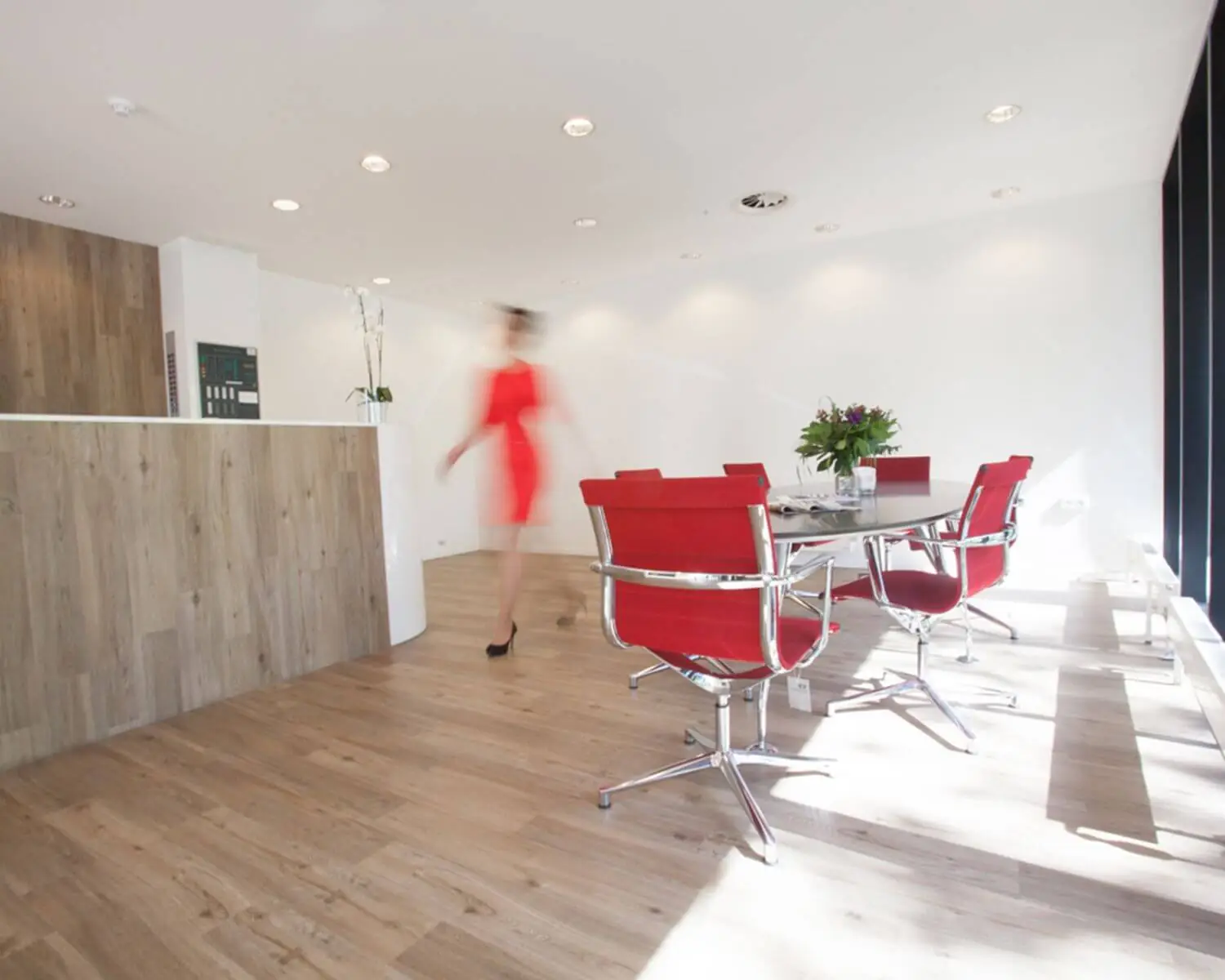 Moderne ontvangstruimte met houten vloer, rode stoelen en een vrouw in een rood jurkje die langs de balie loopt.