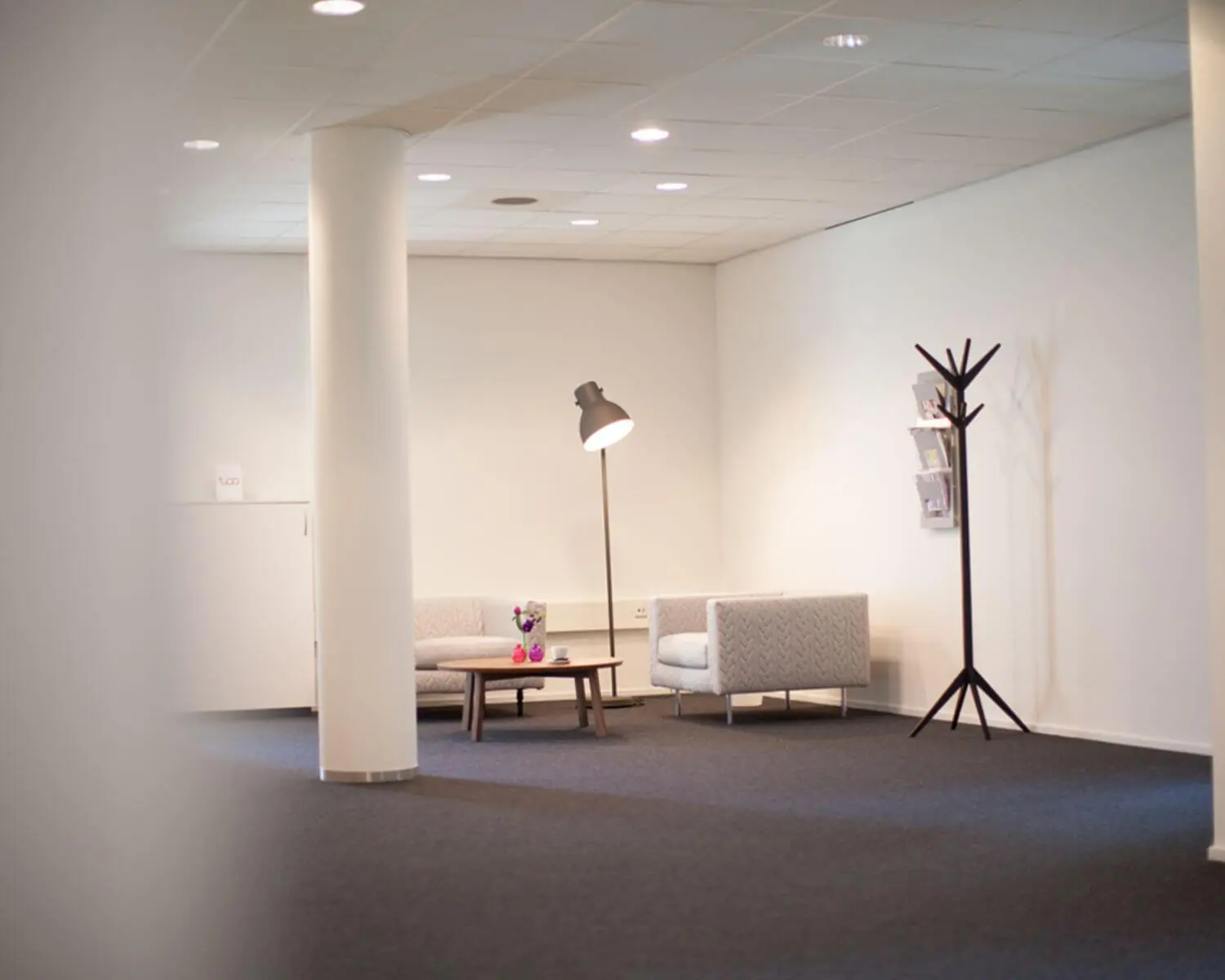 Wachtruimte met twee grijze fauteuils, een staande lamp, kapstok en een tafel met bloemen in een modern kantoorinterieur.