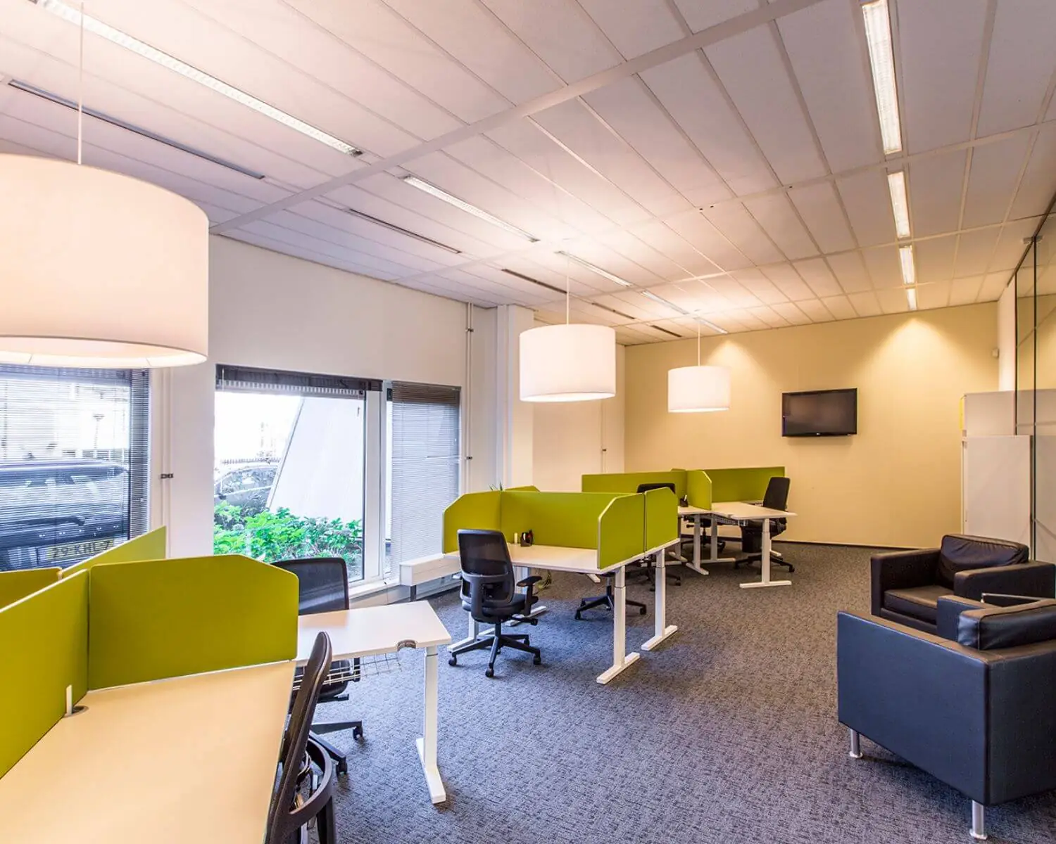 Modern kantoor met open werkplekken, groene scheidingspanelen en een zitgedeelte met zwarte fauteuils.