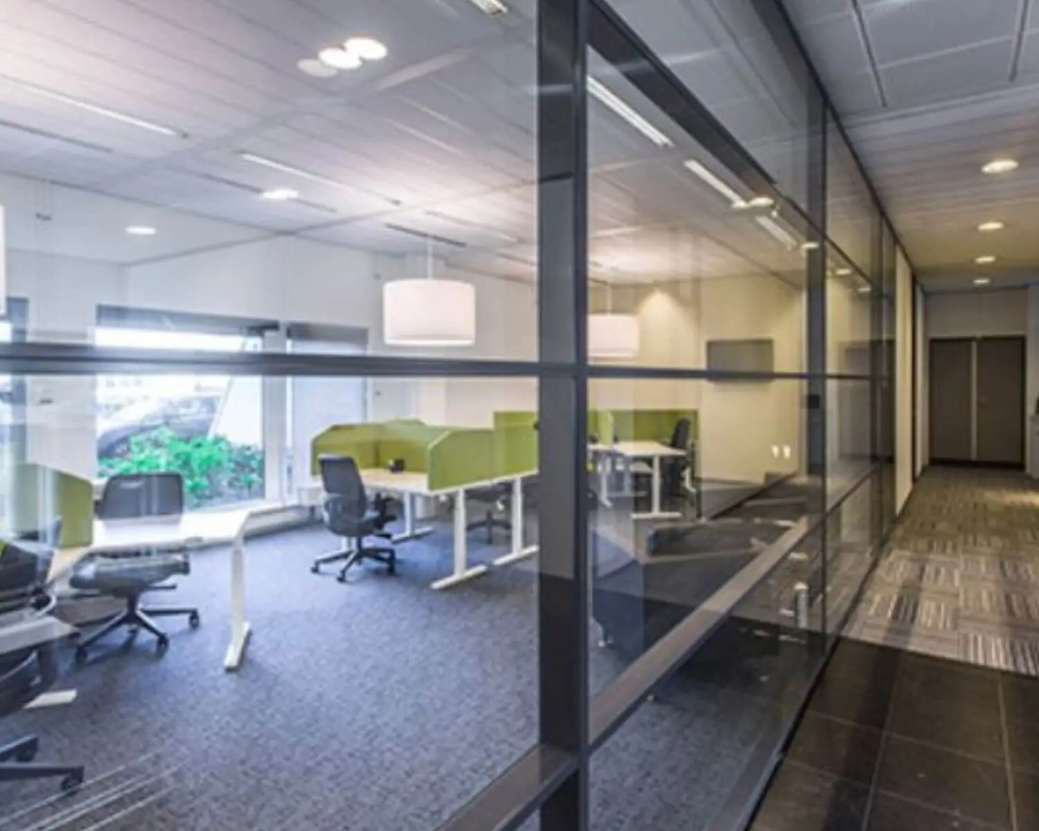 Moderne kantoortuin met glazen wanden, bureaus met groene scheidingspanelen en ergonomische stoelen.