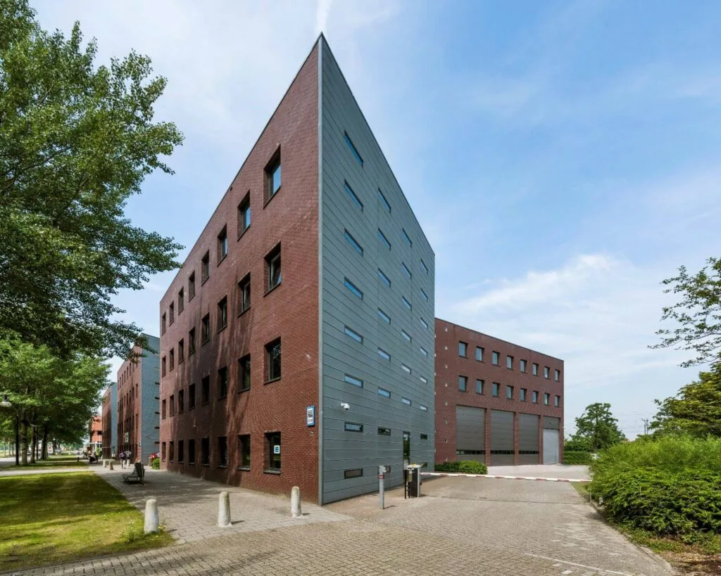 Modern, puntig hoekig gebouw met rode bakstenen en grijze gevelpanelen aan een rustige straat met bomen.