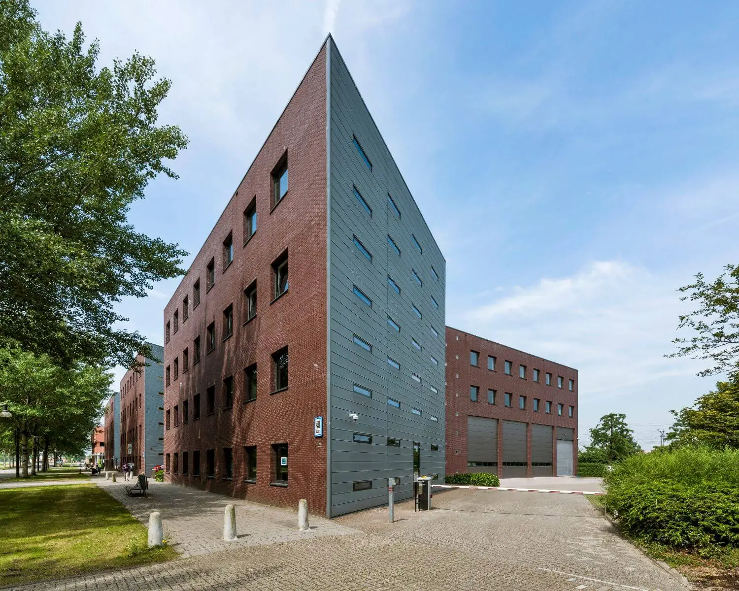 Modern, puntig hoekig gebouw met rode bakstenen en grijze gevelpanelen aan een rustige straat met bomen.
