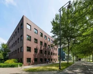 Bakstenen kantoorgebouw met vier verdiepingen aan een boomrijke straat op een zonnige dag.