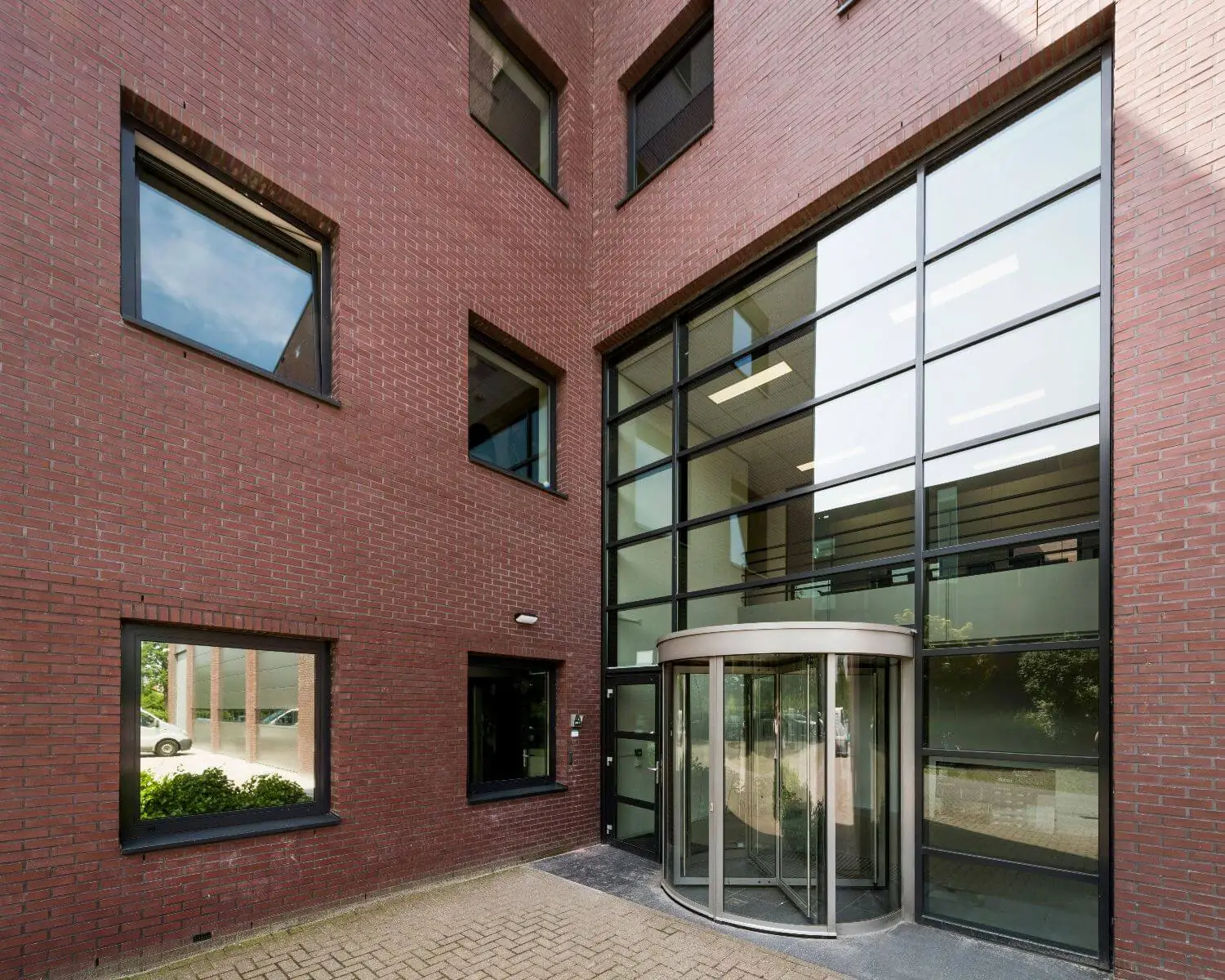 Ingang van een modern kantoorgebouw met een glazen draaideur en grote ramen in een rode bakstenen gevel.