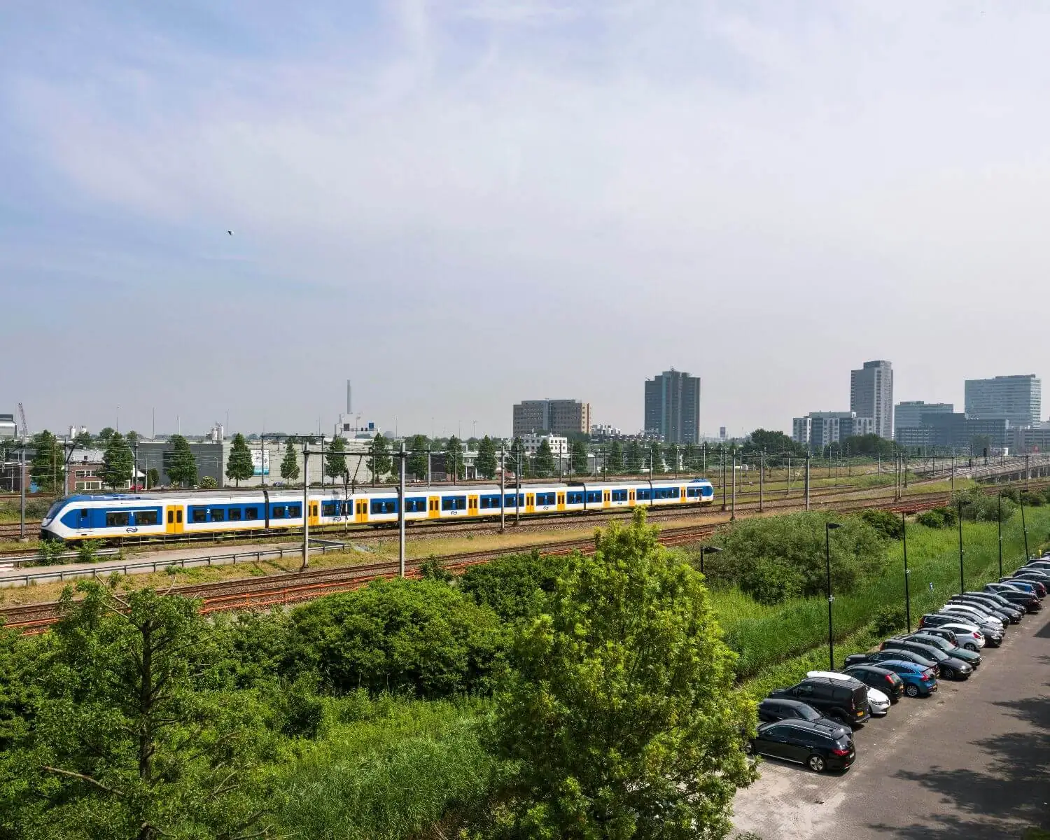 Een NS-sprintertrein rijdt langs een spoorlijn met op de voorgrond een parkeerplaats en op de achtergrond een stedelijke skyline.