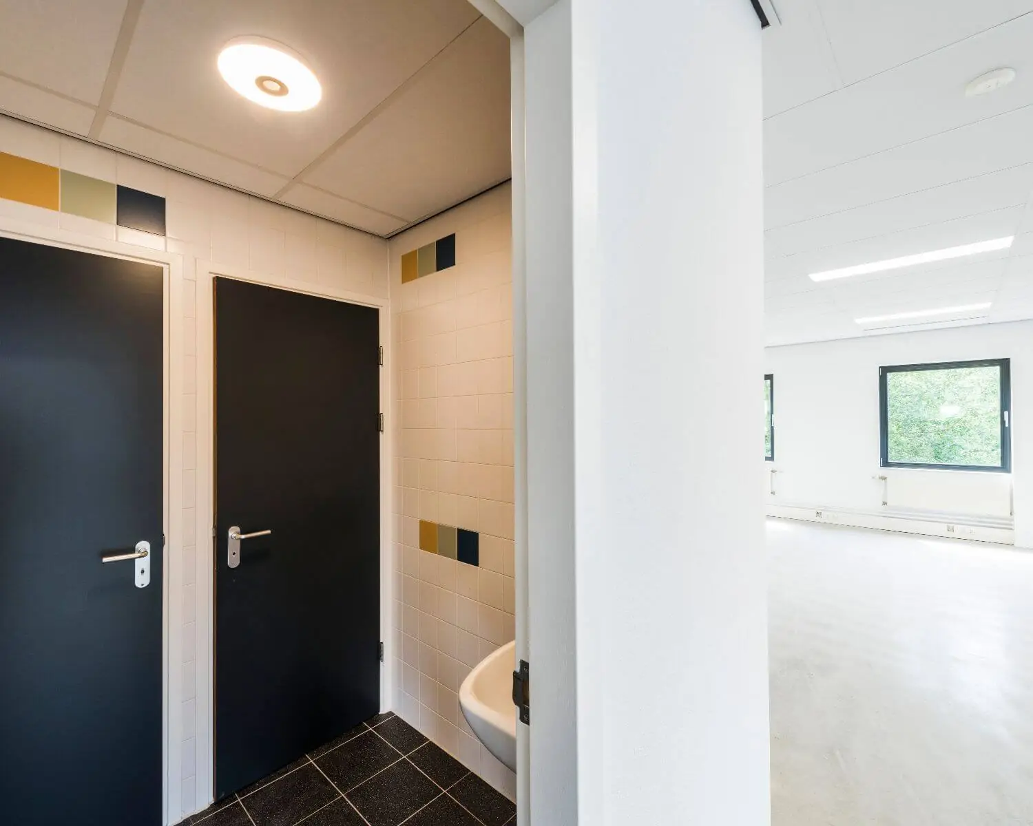 Een nette, lege kantoorruimte met zicht op een toiletruimte met twee zwarte deuren en kleurrijke tegelaccenten.