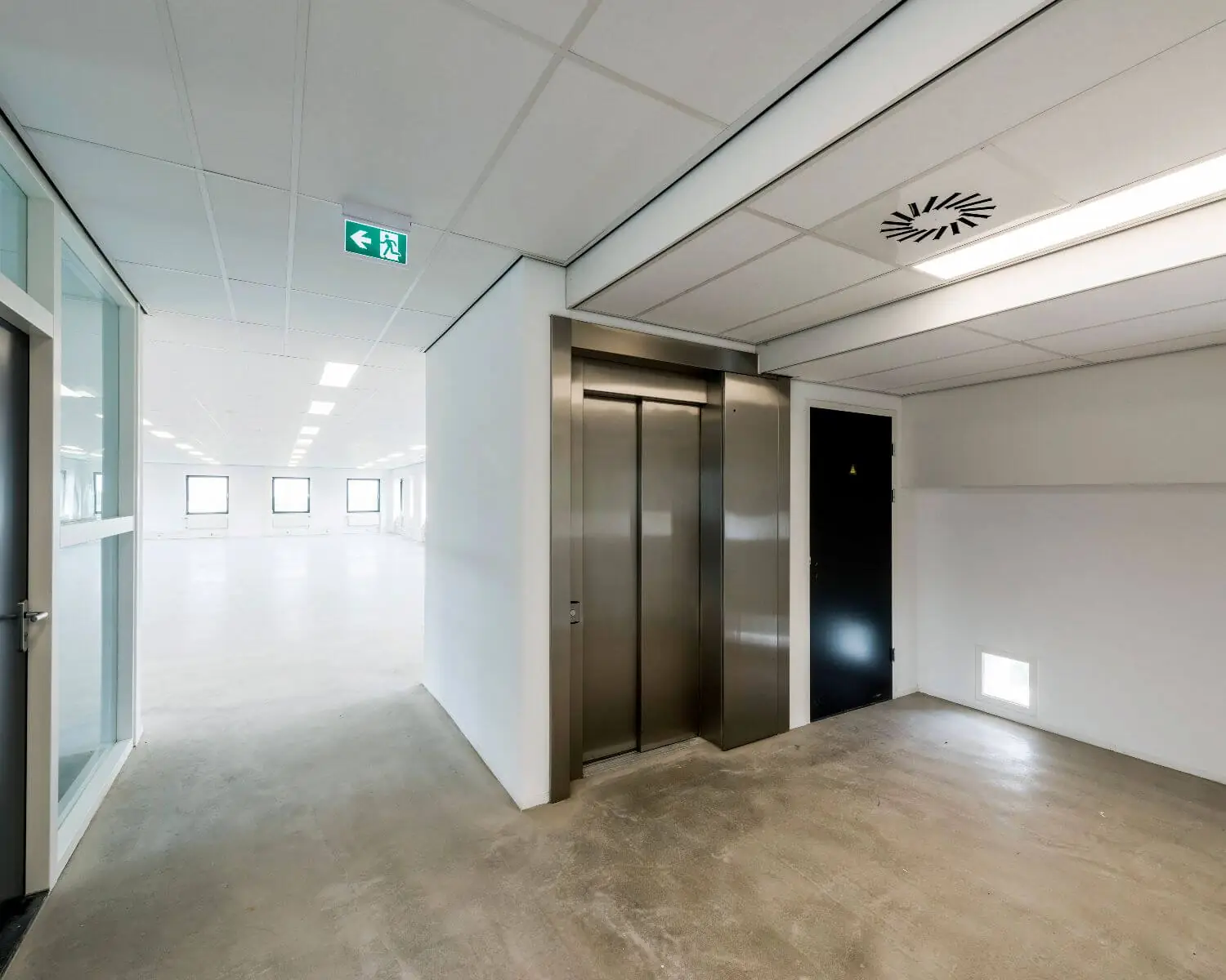 Moderne, lege kantoorruimte met een gesloten liftdeur en nooduitgangsbord aan het plafond.