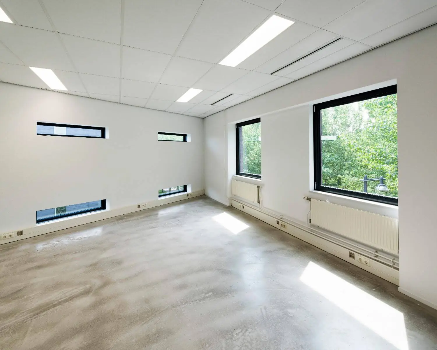 Licht en leeg kantoor met grote ramen, witte muren en betonnen vloer.