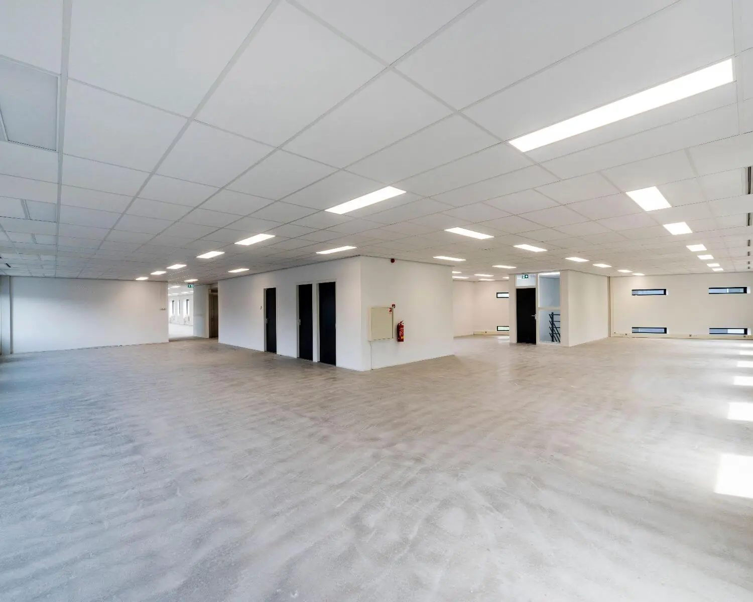 Lege, moderne kantoorruimte met systeemplafond, betonnen vloer en witte muren.