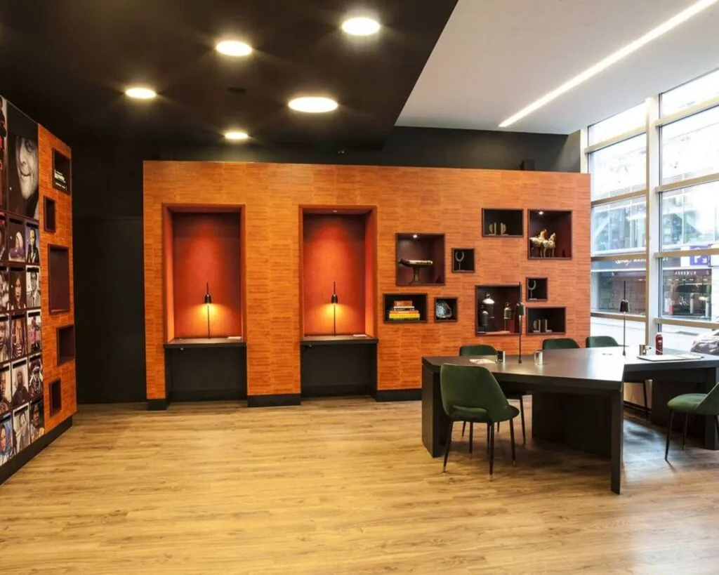 Moderne kantoorruimte met houten vloer, oranje accenten, ingebouwde vitrines en een grote zwarte vergadertafel met groene stoelen.