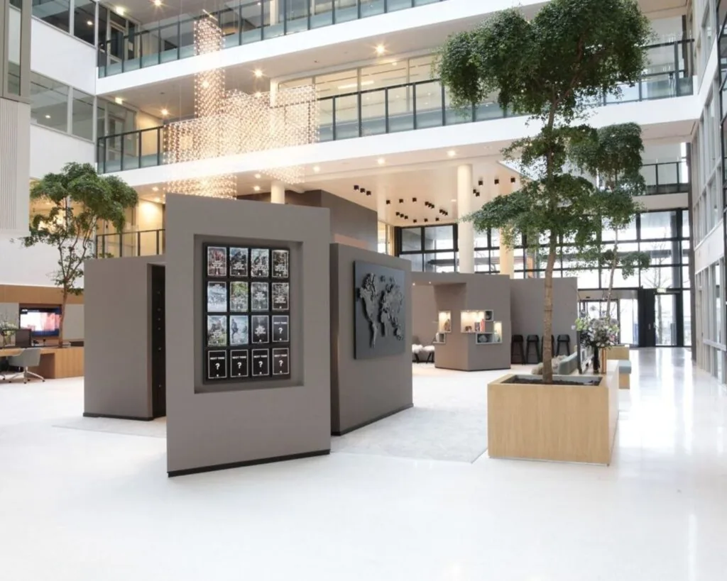 Moderne, open kantoorlobby met veel daglicht, grote plantenbakken en grijze tentoonstellingsstructuren.