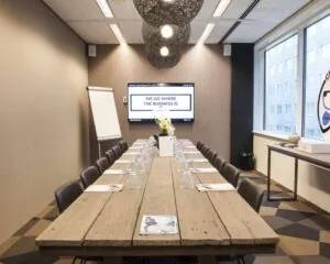 Vergaderruimte met lange houten tafel, zestien stoelen, een scherm met de tekst "We go where the business is", en een flip-over in een moderne kantooromgeving.