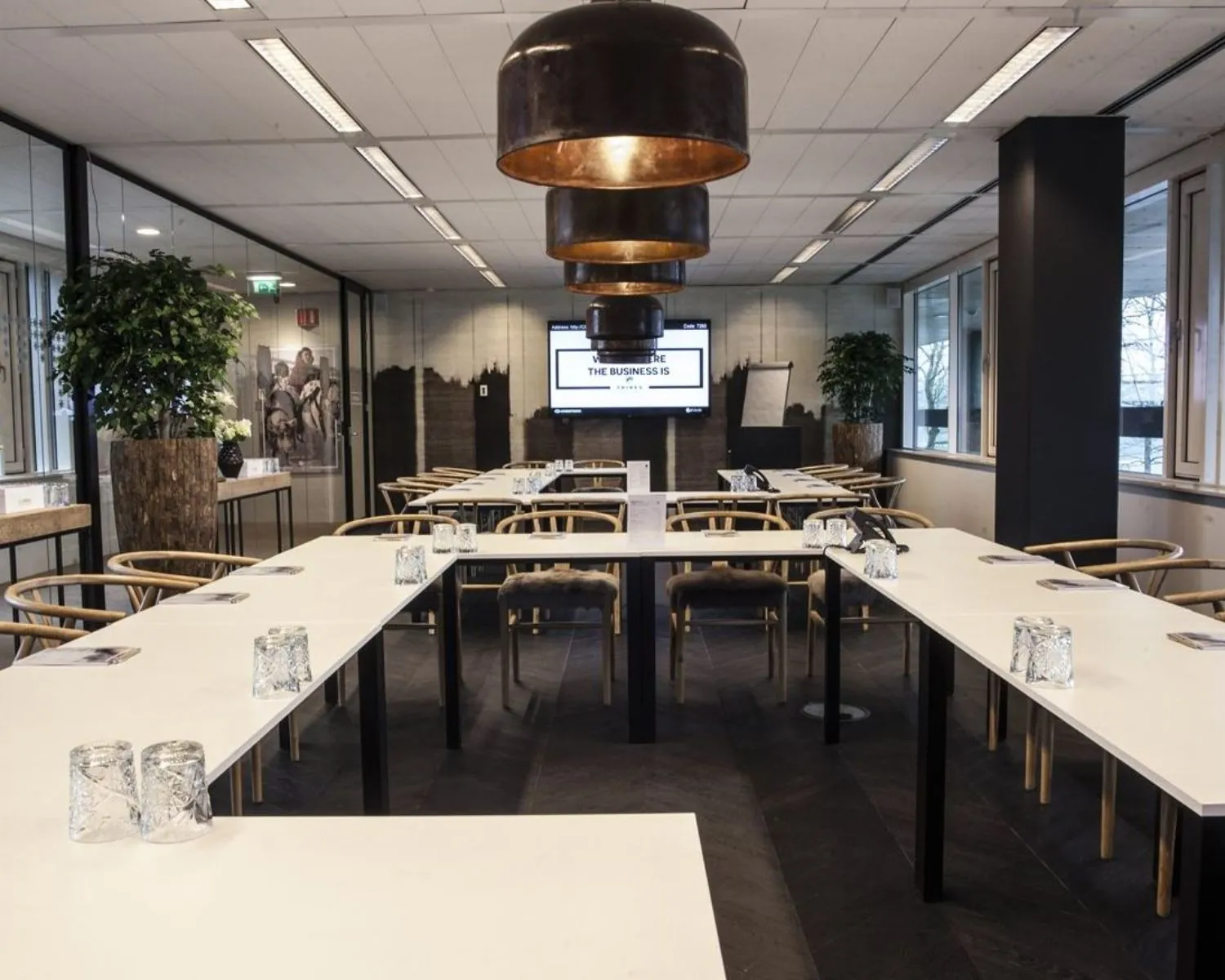 Vergaderruimte met U-vormige opstelling, moderne stoelen, grote hanglampen en een scherm met de tekst "The Business is".