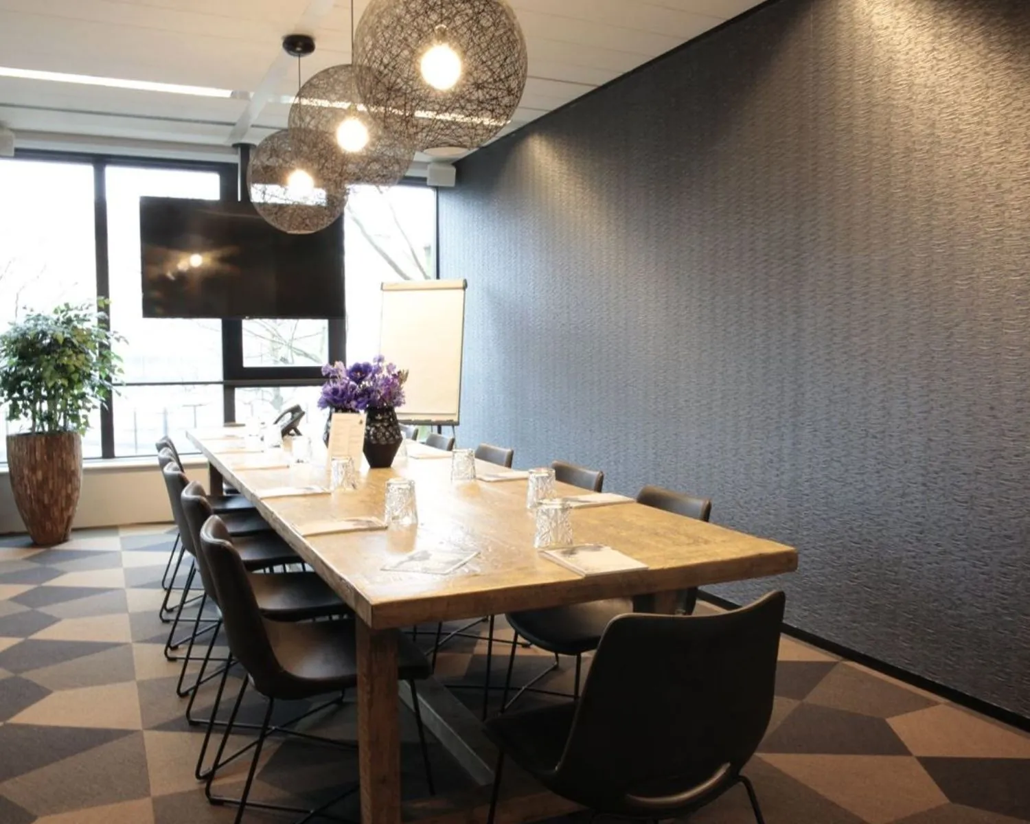 Vergaderruimte met een lange houten tafel, moderne stoelen, een flip-over, grote tv en decoratieve hanglampen.