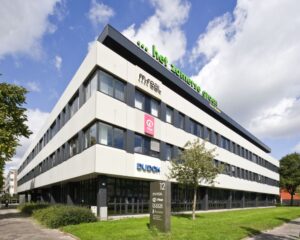 Kantoorgebouw met meerdere bedrijven, waaronder m|real, Plan Nederland en DUDOK, aan een groene straat onder een deels bewolkte lucht.