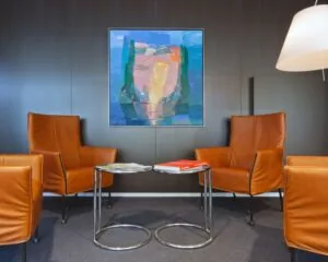 Moderne wachtruimte met vier cognackleurige leren fauteuils, twee ronde bijzettafels en een kleurrijk abstract schilderij aan de muur.