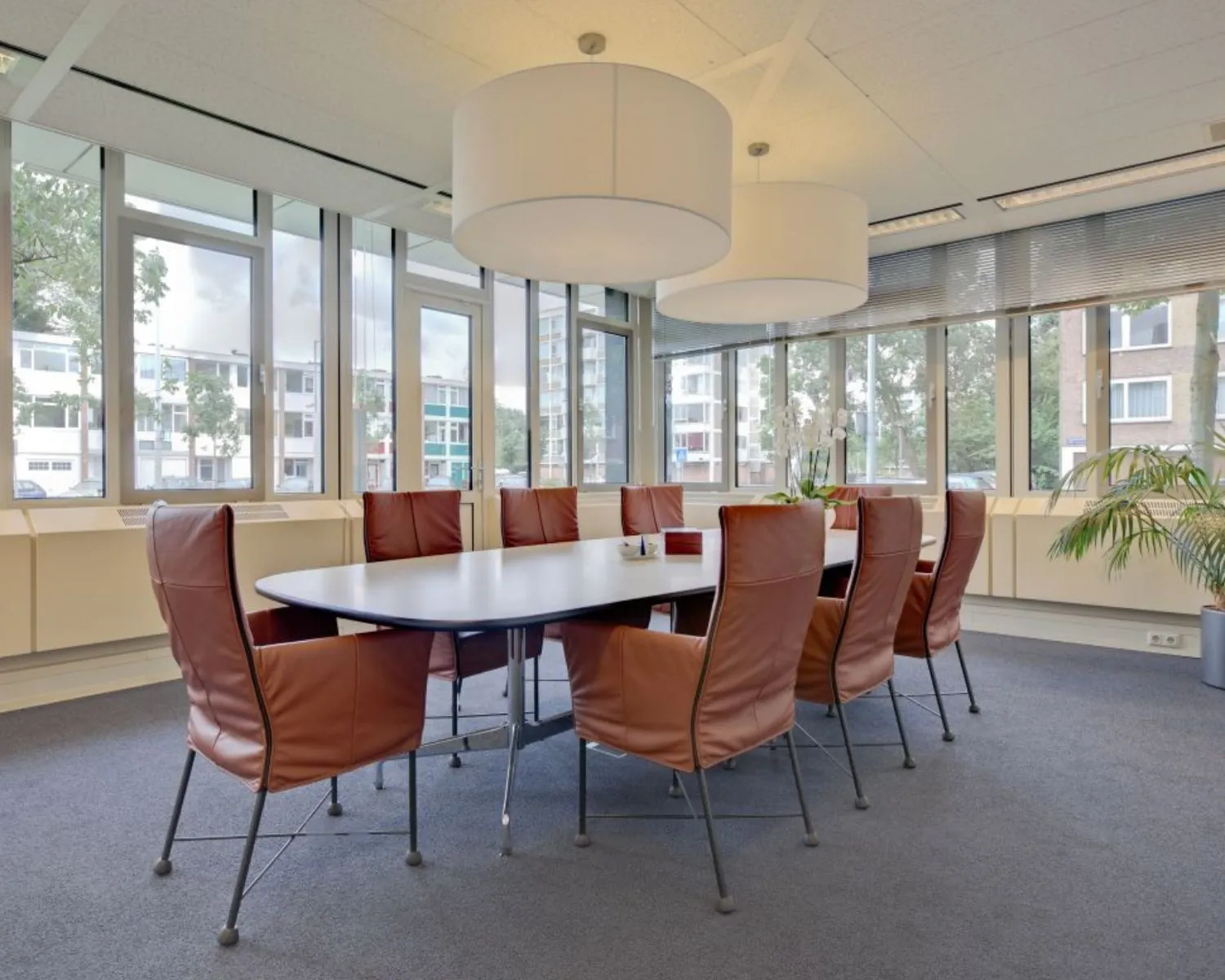 Vergaderruimte met een grote ovale tafel, omringd door bruine leren stoelen en omgeven door ramen.