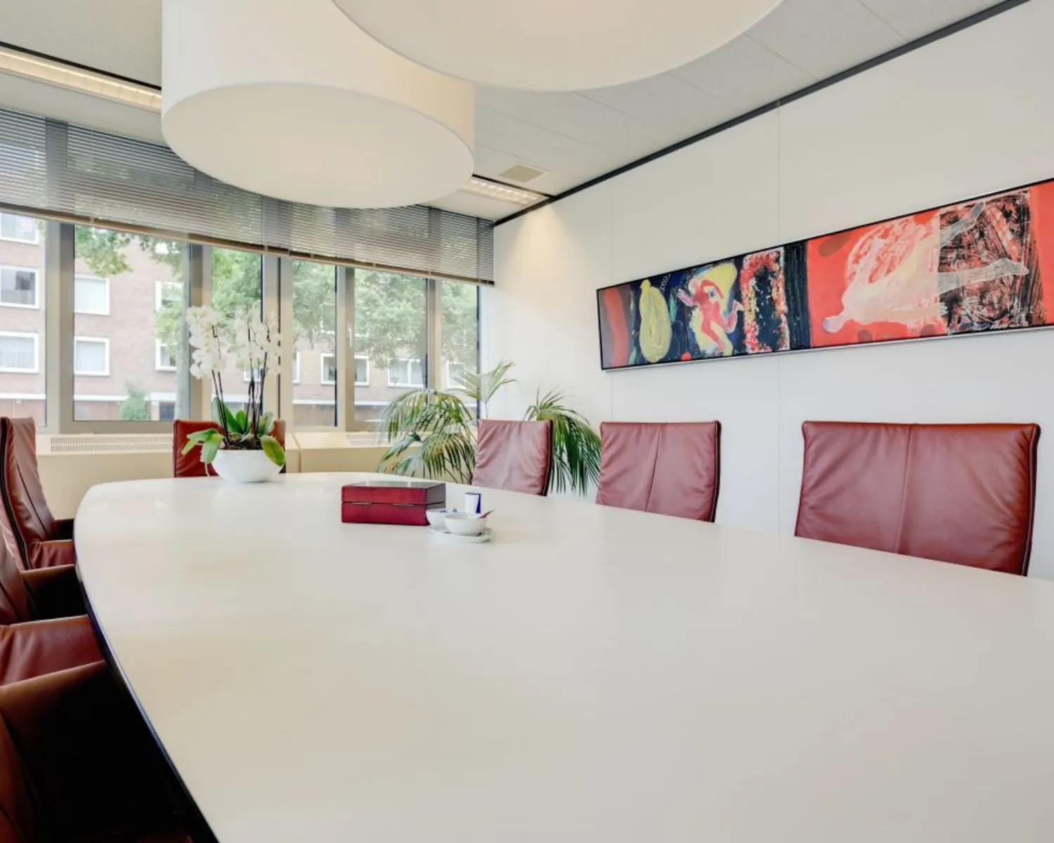Vergaderruimte met lange witte tafel, roodbruine stoelen, grote ramen en kleurrijk modern kunstwerk aan de muur.