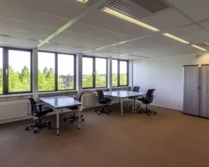 Licht en modern kantoorgedeelte met vier bureaus, bureaustoelen en grote ramen met uitzicht op bomen.