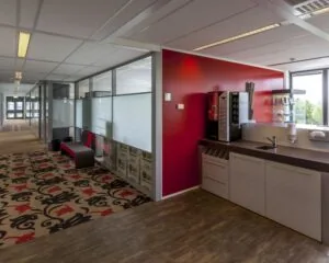 Pantryhoek met koffieautomaat en aanrecht in een modern kantoorpand met felrode muur en kleurrijk tapijt.