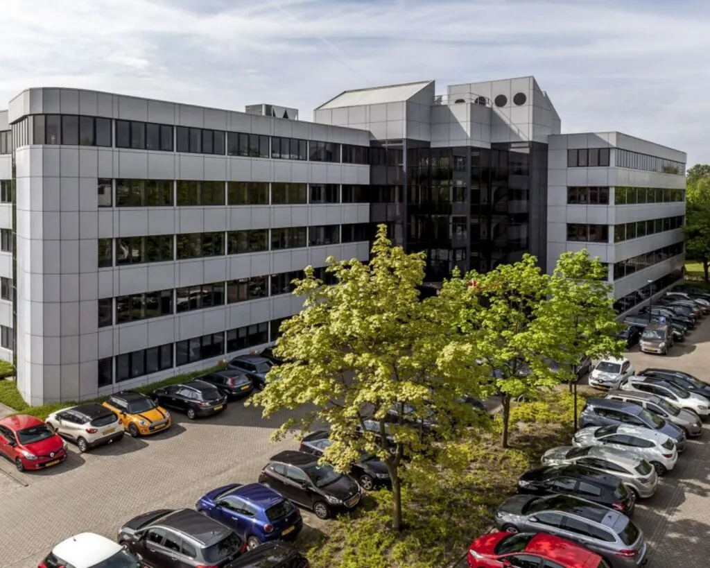 Kantoorgebouw met glazen entree en parkeerplaats vol auto's op de voorgrond.