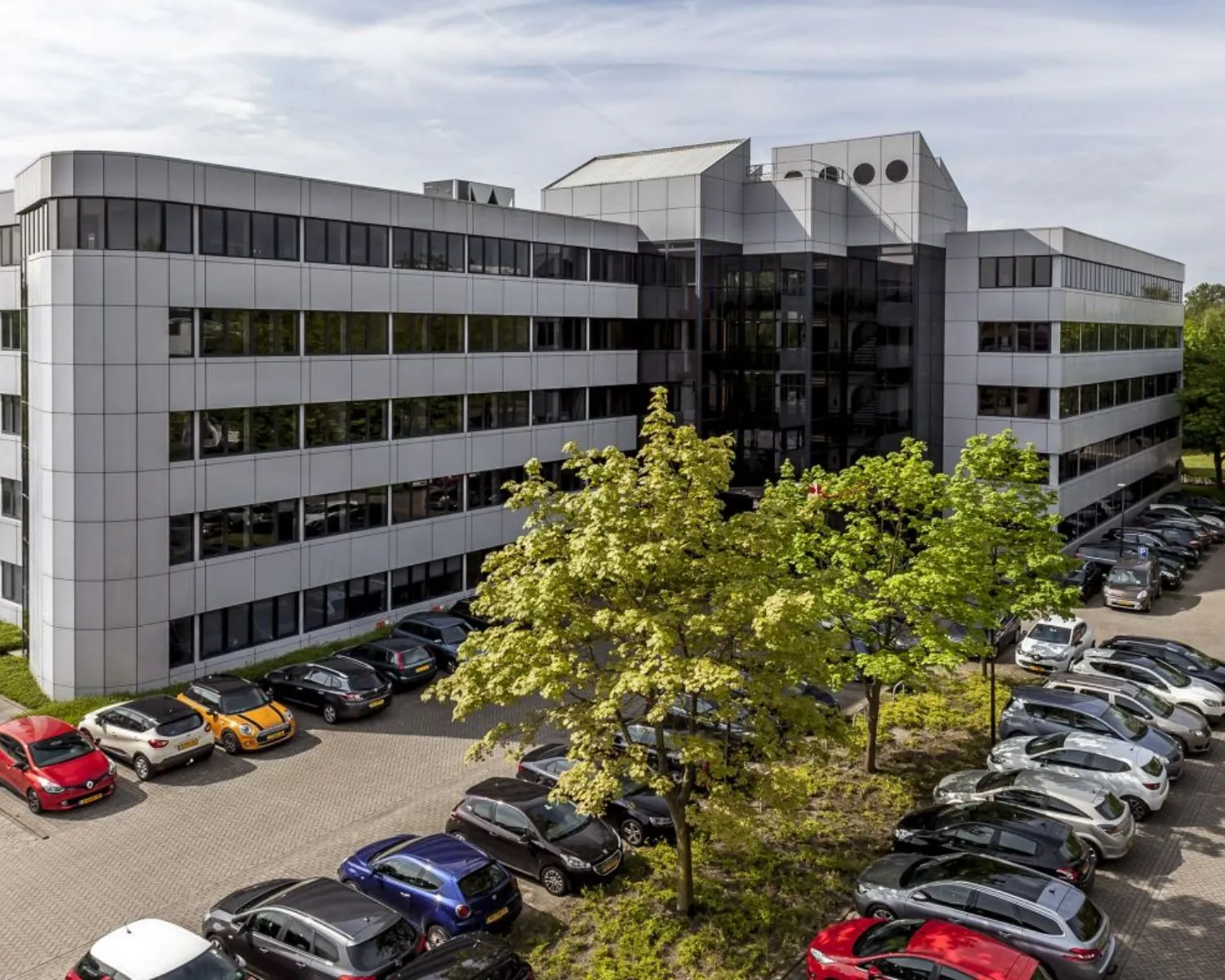 Kantoorgebouw met glazen entree en parkeerplaats vol auto's op de voorgrond.