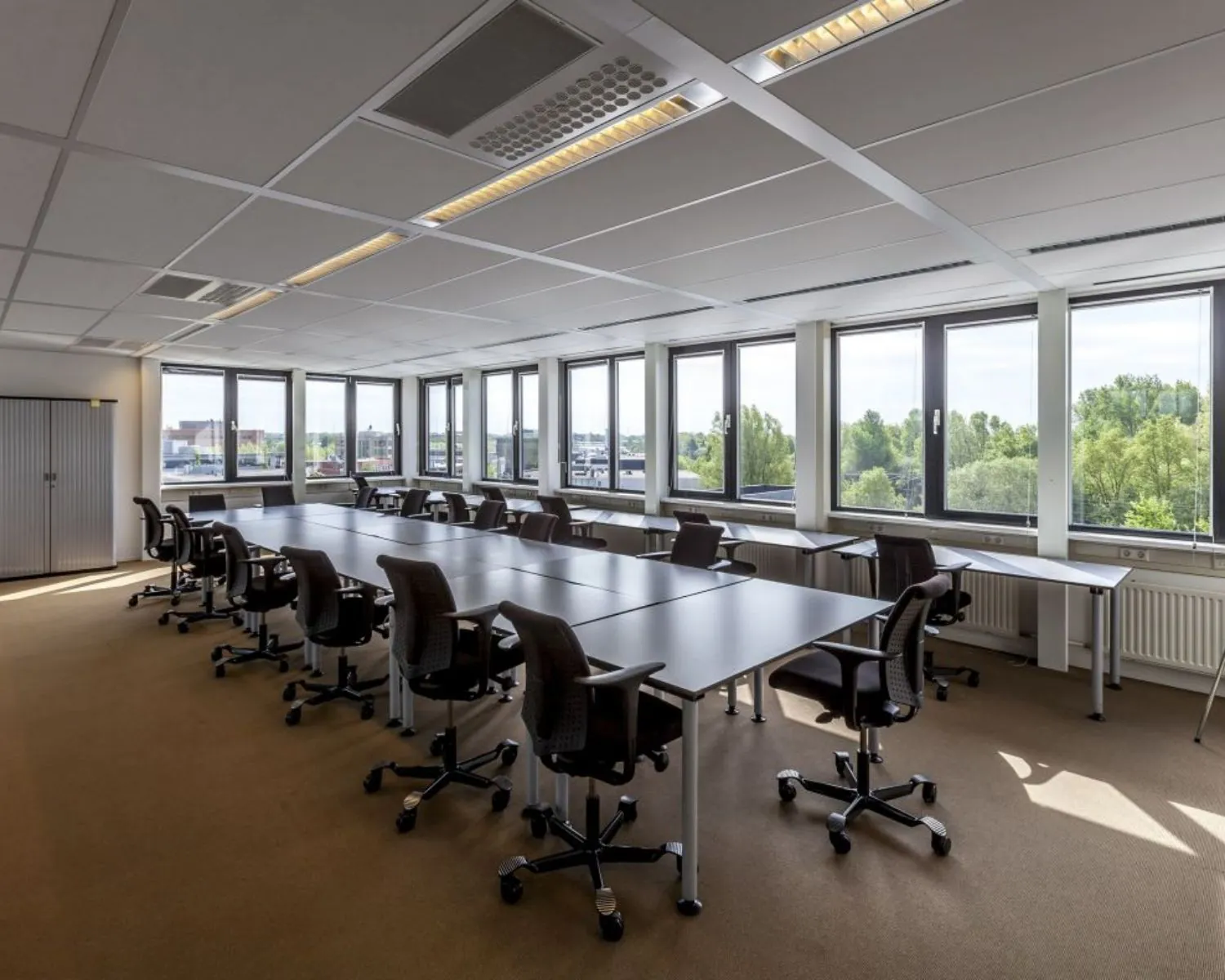 Ruim, modern kantoor met meerdere bureaus en bureaustoelen naast grote ramen met uitzicht op groen.