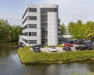 Kantoorgebouw met parkeerplaats aan het water, omgeven door groene bomen.