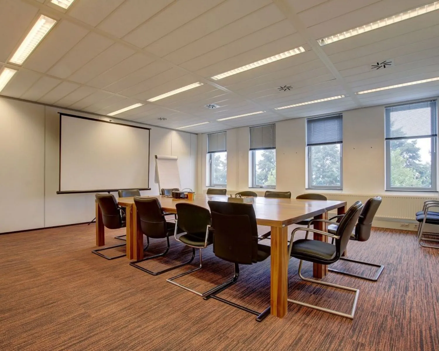Vergaderruimte met een lange houten tafel, zwarte stoelen, een flip-over en een projectiescherm.