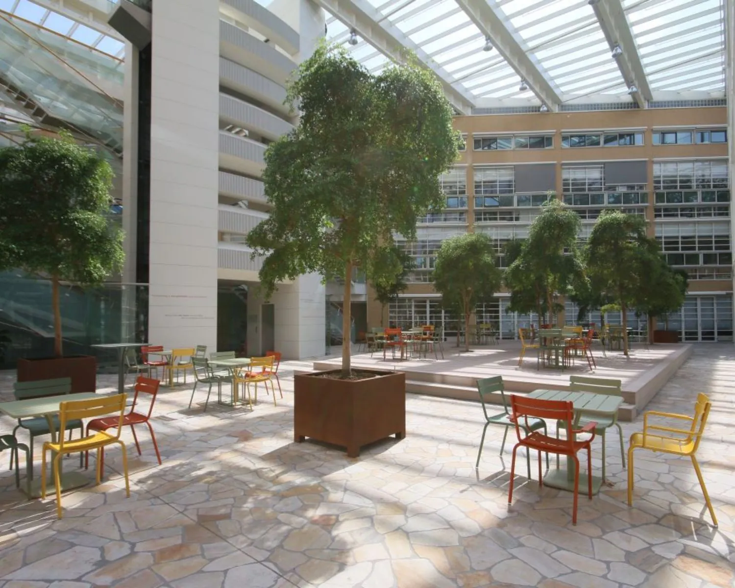 Overdekte binnentuin met kleurrijke tafels en stoelen tussen grote plantenbakken en moderne kantoorgebouwen.