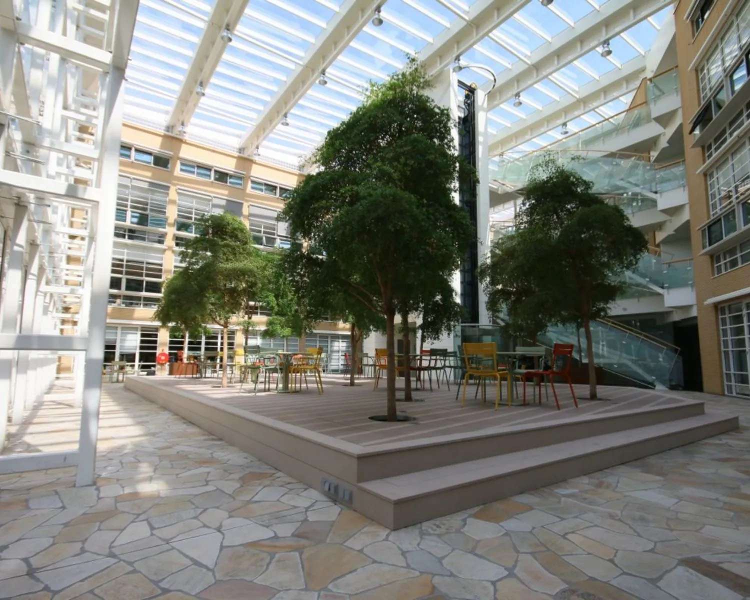Overdekt atrium met bomen, kleurrijke stoelen en tafels op een verhoogd terras in een modern kantoorgebouw.
