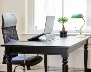 Een zwarte bureautafel met een laptopstandaard, een kamerplant en een groene bureaulamp bij een raam.