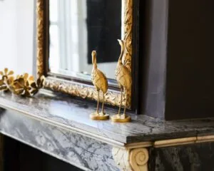 Twee gouden sierlijke vogelbeelden op een marmeren schouw met een grote sierlijstspiegel op de achtergrond.